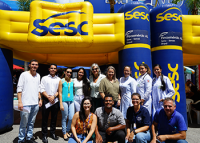 Grupo de mulheres e homen, alguns acocorados, em frente a uma tenda inflável amarela e azul