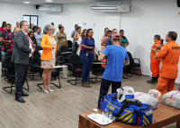 Vários homens e mulheres de pé prestando atenção a fala de dois bombeiros fardados de cor laranja e um homem de camiseta preta e calça preta.