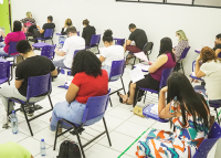 Vários homens e mulheres sentados em carteiras escolares de cor azul escrevendo.