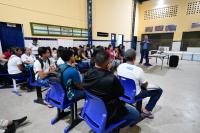 Homem de terno escuro e camisa azul falando para diversas alunos de uniformes brancos sentados em carteiras escolares na cor azul.