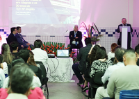 Várias pessoas sentadas ouvindo palestra de um homem de pele clara e terno escuro.
