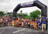 Portal de largada de corrida nas cores azul com letras brancas . Há várias crianças com camisetas laranjas.