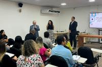 Dois homens de terno e uma mulher da saia estampada e blusa preta em pé na frente de uma plateia composta de homens e mulheres