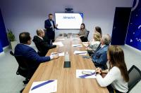 Homem de terno em pé apresentando slide a três homens de terno e quatro mulheres. Todos sentados em uma mesa de madeira