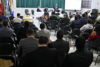 Auditório com várias pessoas sentadas assitindo uma palestra de um homem de pele clara que esta trajando terno escuro. Há dois homens sentados próximas ao palestrante também usando ternos escuros.