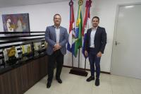 Dois homens de pé na frente de três bandeiras. Ele estão trajando ternos.