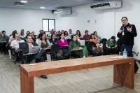 Sala de aula com dezenas de pessoas sentadas, olhando atentamente o professor , que está à frente, usando terno escuro e gesticulando com uma das mãos