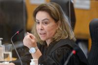 Foto de mulher com pele branca, cabelos castanhos claros na altura dos ombros, vestindo roupa preta e falando ao microfone 