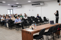 mulher de vestido preto e cabelos pretos dando aula; em segundo plano, vários alunos sentados.