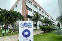 Em primeiro plano, o banner nas cores azul e branco, com um círculo no meio e nele inserido uma seta de indicação na cor branca e, fora a círculo, a frase mutirão vaga inclusiva de trabalho; ao fundo, a fachada da Faculdade Unit.