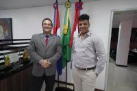 Dois homens morenos. Um tá vestindo terno cinza com gravata veremlha e o outro com roupa clara. Há três bandeiras no local, a Estado de Alagoas, a do Brasil e a do TRT da 19ª Região