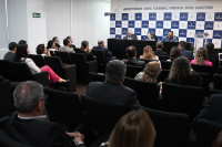 Auditório com homens e mulheres reunidos . Há dois homens de ternos escuros e uma mulher entre eles. Ela veste roupa azul e está falando.