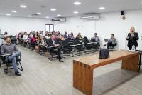Mulher loira, de terno escuro, com microfone na mão à frente de uma turma de pessoas adultas assistindo aula