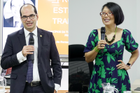 À esquerda homem branco de terno escuro, óculos e calvo, à direita mulher de vestido estampado verde e azul, cabelo preto e curto usando óculos 