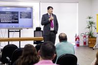 Homem branco, alto, de cabelo preto, usando paletó preto e gravata rosa,  em pé ministrando palestra em um auditório; ao fundo, a imagem de um telão com slides em uma caixa com fundo azul e letras brancas. 