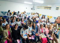 Plateia com vários alunos uniformizados de camisetas brncas e calças jeans. Há  várias mulheres com vestidos de diversas cores.