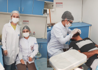 Profissionais de saúde fazem atendimento odontológico em homem de barba deitado em cadeira de dentista
