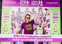Mulher branca de óculos escuros vestindo uma camisa roxa com nomes amarelos-limões, ao fundo, um backdrop com as fotos de vários atletas correndo