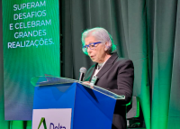 Mulher branca de cabelos brancos de pé falando em um púlpido azul. Ela está de óculos azuis e roupa escura. Atrás dela há projeção de luz verde nas cortinas.