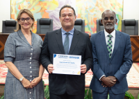 Há três pessoas de pé posando para foto. Sendo uma mulher de vestido cinza e dois homens trajando ternos escuros. O homen que está posicionado no meio segura um certificado de premiação.