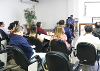 Foto de um auditório com servidores sentados de costas voltados para a frente do auditório; de frente para os servidores, um palestrante sentado na cadeira ao lado do birô vestindo camisa azul clara e calça preta