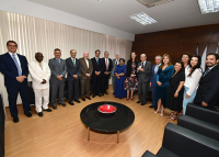 Vários homens e mulheres em pé reunidos para uma fotografia. Eles estão em uma sala com piso claro. Há no meio do local uma mesa de centro preta com um adorno vermelho.