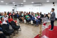 Várias pessoas sentadas assistindo uma palestra. Há um púlpito de acrílico em destaque  ao lado de um homem de calça escura e blese cinza claro. Ele está com um micrpfone na mão falando para o público.