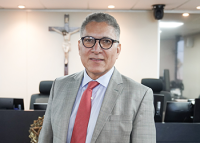 Foto de meio busto de homem pardo de cabelos curtos brancos usando óculos preto redondo, terno cinza, gravata vermelha e blusa branca