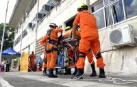 Bombeiros com uniformes na cor laranja levando uma maca. Eles simulam um socorro.