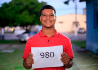 Jovem negro, usando camiseta laranja, segura sorridente uma foloha de papel com o número 980