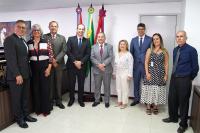 Grupo de homens e mulheres posando para fotos. Ele estão em uma sala branca, na qual há três bandeiras, sendo a do Brasil localizada ao centro.