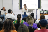 Mulher branca de cabelos claros, vestida de branco e azul falando para uma plateiaa. Há também uma mulher de pele clara sentada ao lada da que fala.