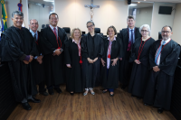 Grupo de nove pessoas, todas vestindo togas pretas, sendo que ao centro, sorridente, a juíza do Trabalho Ana Cristina, ladeada pelas desembargadoras Vanda e Eliane, e ao lado delas, o juiz Alonso, os desembargadores Catão, João Leite, Anne e Laerte, além do procurador Rafael Gazzaneo