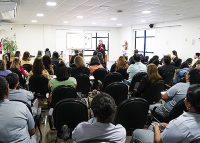Várias pessoas sentadas assistindo ua palestra. Há uma mulher de roupa preta e vermelha de pé falando para plateia.