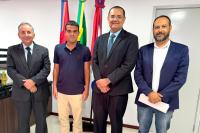 Quatro homens perfilados e sorridentes posam para foto em frente às bandeiras do Estado, TRT e Brasil. Três deles vestem terno e o mais jovem, camisa polo azul.