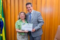 Mulher branca de meia idade, vestida de blusa verde e saia cinza com estampas coloridas. Ela está ao lado de um homem também branco que veste terno cinza claro. Eles seguram um certificado.