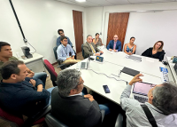 Vários homnes e mulheres sentados. Eles estão reunidos em volta de uma mesa branca.