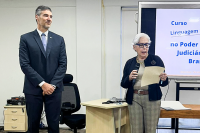 Homem alto de paletó azul-marinho escuro à esquerda; à direita, mulher branca de cabelo grisalho lendo com uma folha de papel na mão esquerda.