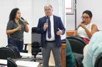 À esquerda, mulher morena, baixa, de cabelo preto em pé com um microfone na mão esquerda; ao centro, um homem  em pé, calvo, moreno, de óculos, paletó azul-marinho, gravata azul marinho e blusa branca; à direita, uma mulher morena, sentada, de cabelo preto e curto, usando blusa branca 
