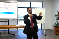 Homem moreno, em pé, de cabelo preto, usando óculos e paletó preto, camisa branca, gravata vinho e com um microfone na mão direita