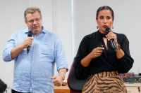 Homem branco de óculos com camisa de manga comprida azul. Há uma mulher de pele clara vestida de blusa de manga comprida preta e saia bege com listras horizontais na cor preta. Os dois têm seguram microfones.