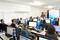 Vários servidores sentados, de costas,  à frente de seus computadores; à frente, homem de altura mediana, cabelo preto, em pé lecionando a aula, usando camisa de cor roxa.