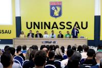 Público sentado assitindo palestra. Há uma mesa grande composta de várias pessoas. Há um homem de terno escuro de pé. Ele está falando ao microfone.