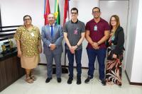 Três homens e duas mulheres posando para uma foto. Eles estão de pé. As pessoas estão na frete das bandeiras do Brasil, de Alagoas e do TRT-19.