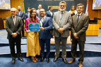 Da esquerda à direita, todos em pé, homem moreno, calvo, usando terno verde escuro; mulher usando saia longa amarela e blusa rosa choque. Essa mulher está segurando um certificado azul com um homem baixo, calvo, que veste terno azul-claro; ao lado dele, homem alto, de cabelos grisalhos utilizando terno bege claro e homem usando óculos que utiliza um terno marrom escuro