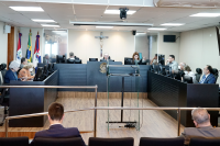 Sala de audiência com homens e mulheres sentados atrás de bancadas com computadores e um púlpito transparente no meio do palco