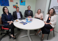 Dois homens de terno e três mulheres em trajes sociais sentados em volta de uma mesa redonda