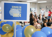 À esquerda, placa com moldura branca e o fundo azul, com o nome Semana da Conciliação Trabalhista 2024 escrito em letra branca; abaixo, da placa, várias bolas se sopro, nas cores azul e amarela e, ao fundo, várias pessoas sentadas esperando atendimento.