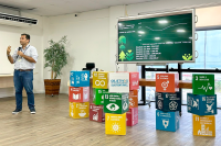 Homem de pé faz apresentação ao lado de caixas coloridas que simbolizam os Objetivos de Desenvolovimento Sustentável da ONU