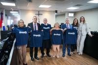 Na foto, em pé, cinco pessoas, sendo três homens e duas mulhers, usando segurando blusas azuis-marinho com o nome empatia escrito em letra branca; ao lado direito deles, duas mulheres: uma com a referida blusa azul-marinho e outra de calça branca, blusa branca e casaco branco.